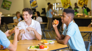 3 jongens aan tafel in de schoolkantine. 2 zijn er aan het eten, eentje niet.